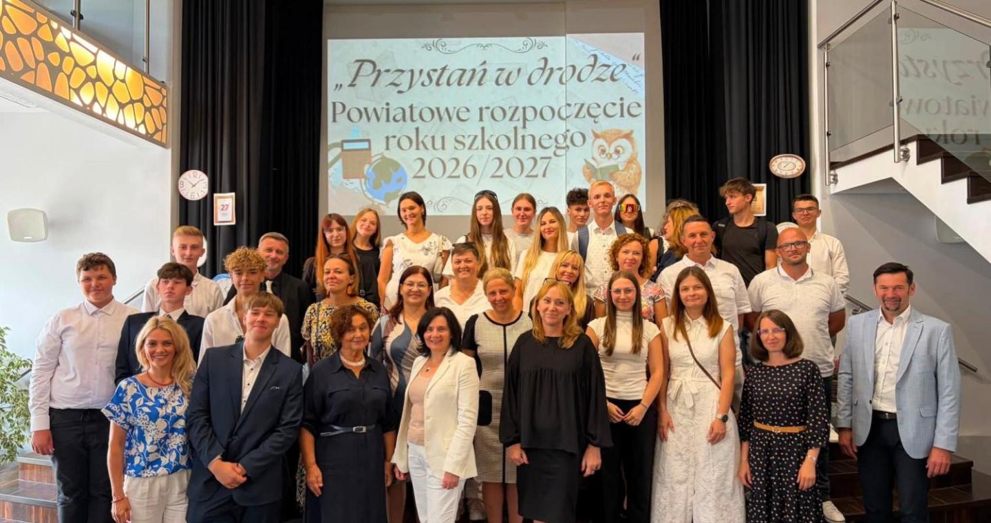 Grupa uczniów i dorosłych pozuje do wspólnego zdjęcia na schodach przed sceną. W tle wyświetlana jest prezentacja dotycząca powiatowego rozpoczęcia roku szkolnego 2026/2027. Po bokach sceny widoczne są zegary oraz kartki z cyframi.