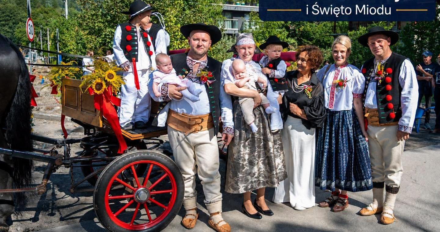 Grupa dorosłych i dzieci pozuje do wspólnego zdjęcia obok udekorowanego wozu zaprzęgowego. Część osób ma na sobie tradycyjne stroje regionalne, a niektórzy trzymają dzieci na rękach. W tle widoczne są drzewa, zabudowania oraz napis „Wiślańskie Dożynki i Święto Miodu”.
