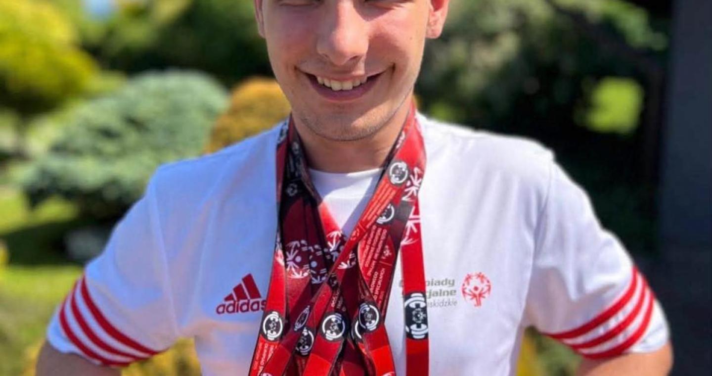 Szymon Machej stoi na zewnątrz w koszulce Olimpiad Specjalnych, uśmiecha się i prezentuje zawieszone na szyi medale zdobyte podczas zawodów.