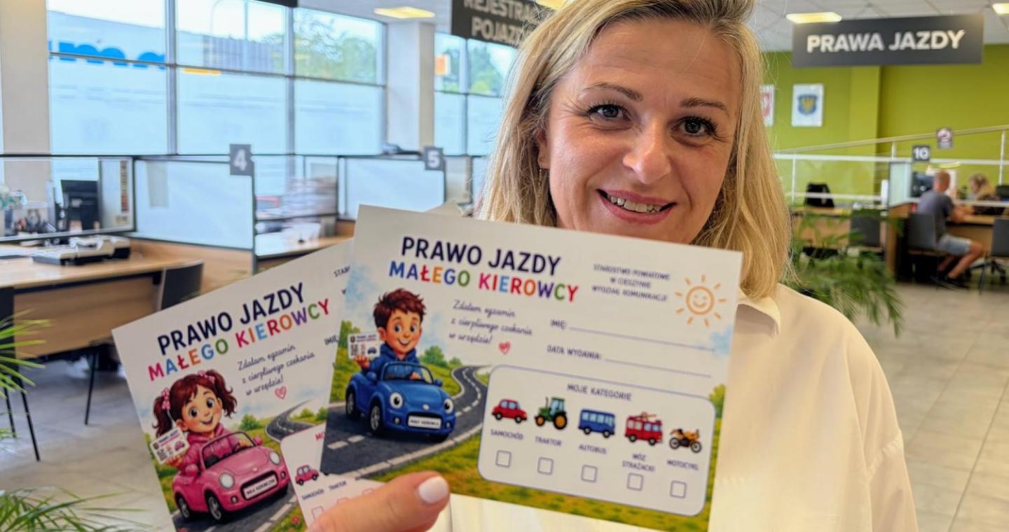 Kobieta trzyma w dłoni materiały zatytułowane „Prawo jazdy małego kierowcy”, ozdobione ilustracjami dzieci i kolorowych samochodów. W tle widoczne są stanowiska obsługi, duże okna oraz tablice z napisami „Rejestracja pojazdów” i „Prawa jazdy”.