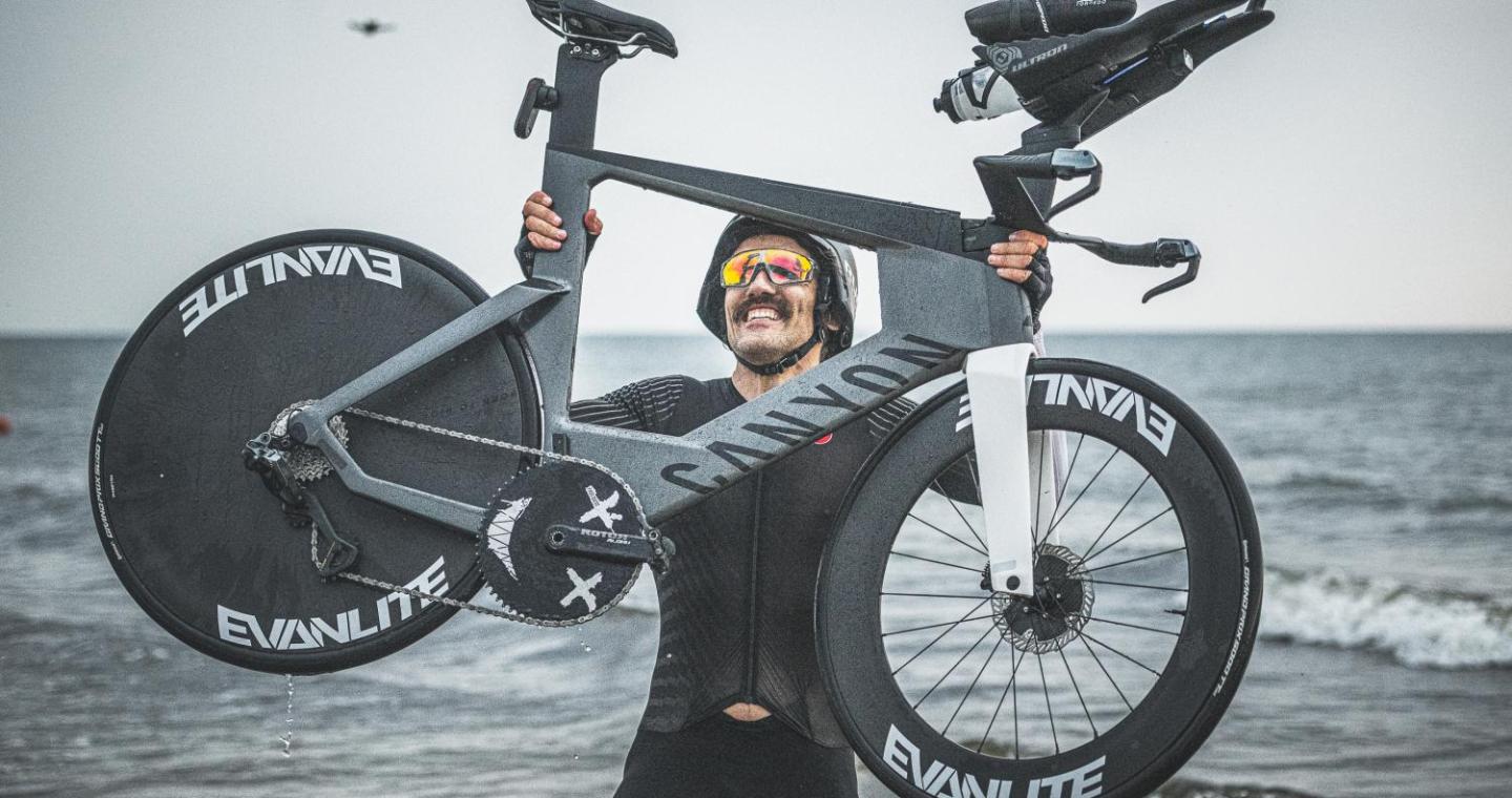 Mężczyzna w stroju sportowym i kasku unosi nad głową rower, stojąc w płytkiej wodzie przy brzegu morza. Ma na sobie sportowe okulary, a na twarzy widoczny jest uśmiech. W tle rozciąga się morze i pochmurne niebo.