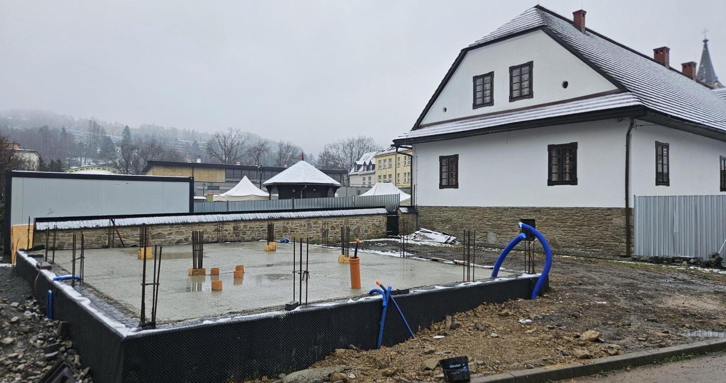 Plac budowy z wykonaną betonową płytą fundamentową i wystającym zbrojeniem, obok zabytkowego budynku w zimowej scenerii.