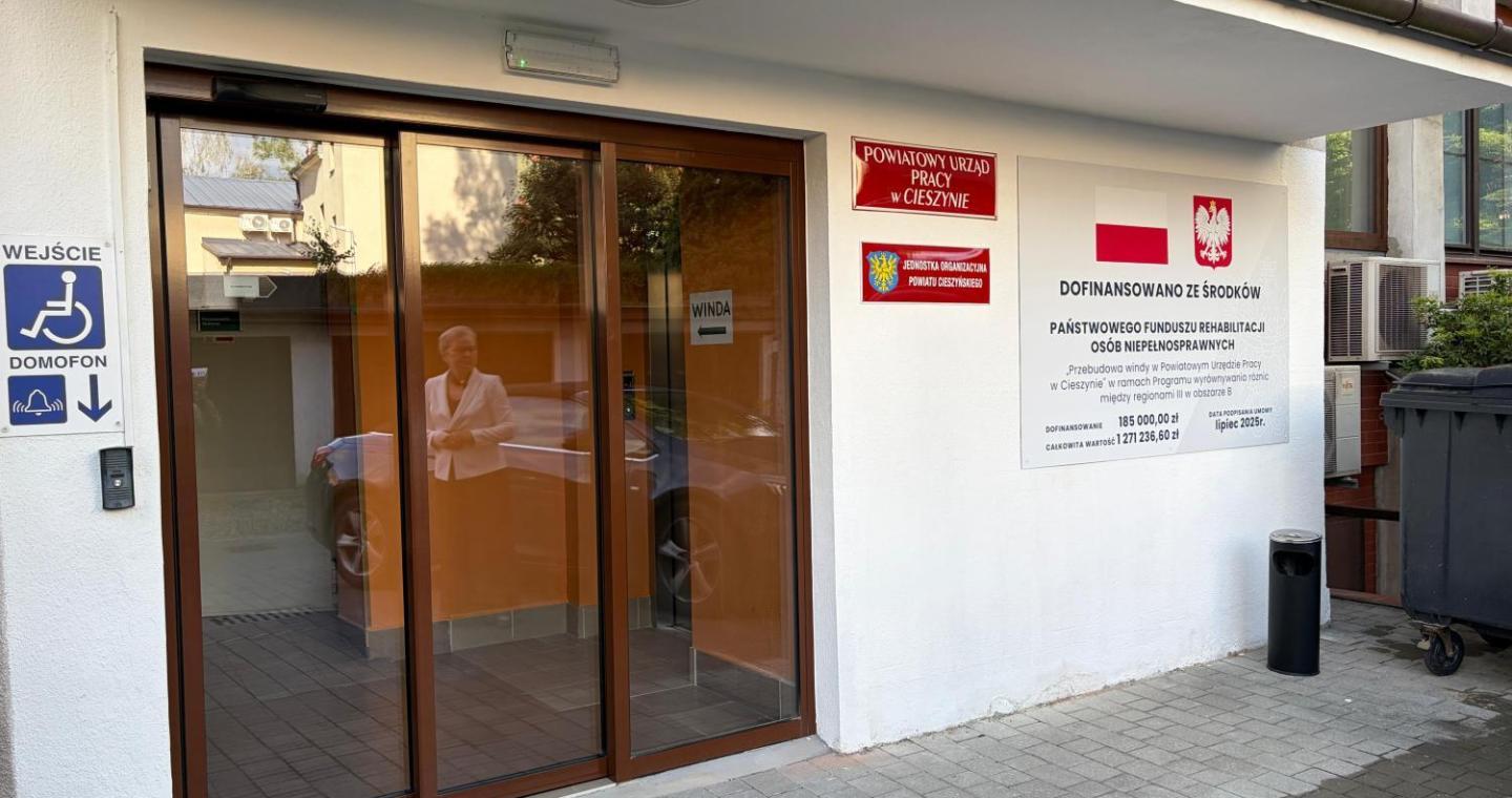 Kobieta prezentuje windę, w której stoją dwie osoby; obok znak „WINDA” ze strzałką w lewo.