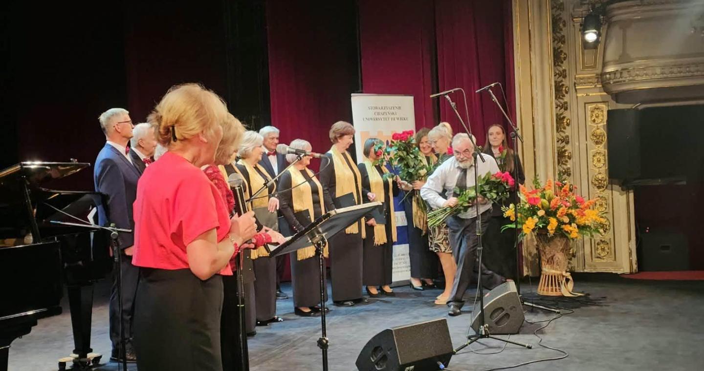 Uroczysta inauguracja Uniwersytetu Trzeciego Wieku w Cieszynie: uczestnicy stoją na scenie, a mężczyzna wręcza kobiecie bukiet kwiatów.