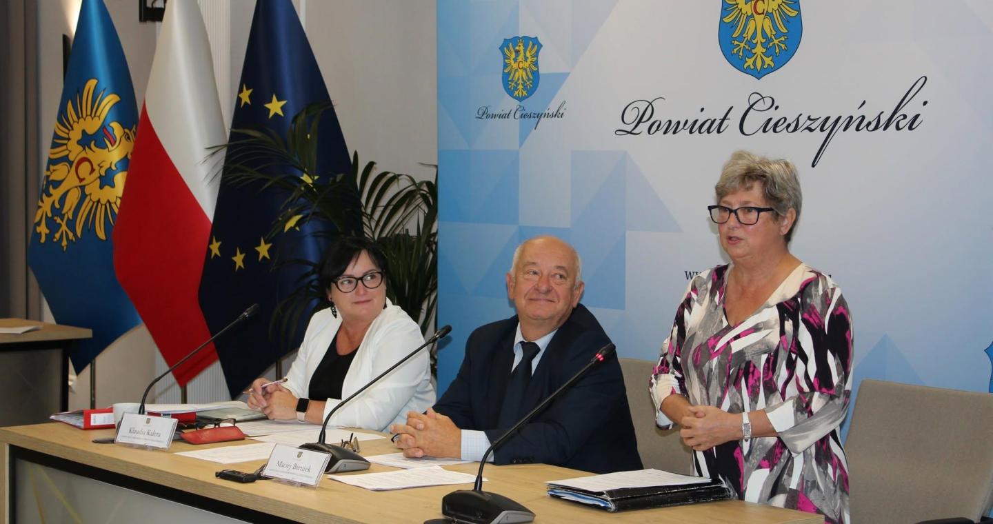 Uczestnicy VIII sesji Rady Seniorów Powiatu Cieszyńskiego przy stole obrad; kobieta przemawia, za nimi flagi i herb powiatu.