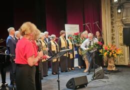 Uroczysta inauguracja Uniwersytetu Trzeciego Wieku w Cieszynie: uczestnicy stoją na scenie, a mężczyzna wręcza kobiecie bukiet kwiatów.