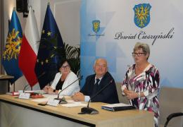 Uczestnicy VIII sesji Rady Seniorów Powiatu Cieszyńskiego przy stole obrad; kobieta przemawia, za nimi flagi i herb powiatu.
