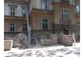 Roboty budowlane przy elewacji budynku; widoczne palety z cegłami, gruz, zsyp z okna oraz siedzący pracownik.