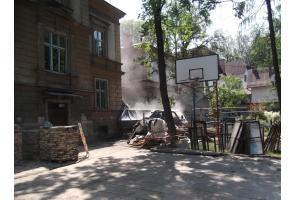 Teren przy budynku w trakcie remontu; widoczne palety z cegłami, ramy okienne, kosz do koszykówki oraz kurz unoszący się w powietrzu.