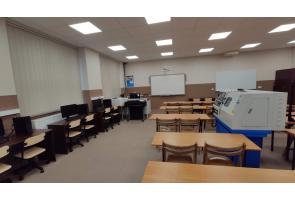 Sala dydaktyczna z rzutnikiem, tablicą, rzędami ławek oraz komputerami po lewej stronie; po prawej urządzenie techniczne, prawdopodobnie tokarka CNC - sala w ZST w Ustroniu.