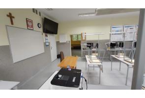 Sala lekcyjna z białą tablicą, projektorem, biurkiem nauczyciela i metalowymi stanowiskami uczniowskimi; na ścianie krzyż, zegar i tablice informacyjne - sala w ZSTiO w Skoczowie.