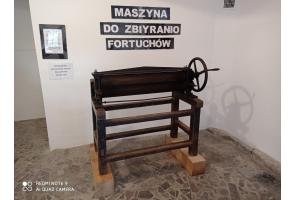 Zabytkowa maszyna z metalowym walcem i korbą, osadzona na drewnianej ramie. Ustawiona na kamiennej podłodze przy białej ścianie. Nad nią napis: „Maszyna do zbiyranio fortuchów”. Obok tabliczka informacyjna.