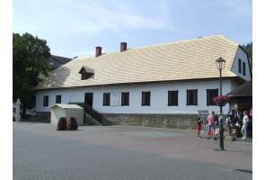 Budynek z białą elewacją i nowym jasnym dachem pokrytym gontem. Przed wejściem schody i daszek. W tle drzewa. Po prawej stronie grupka osób i latarnia uliczna. Na placu brukowana nawierzchnia.