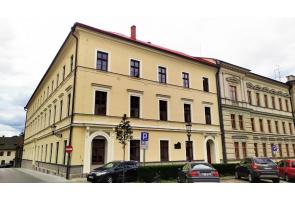 Zabytkowy budynek szkoły Zespołu Szkół Budowlanych  w Cieszynie z jasną elewacją i czerwonym dachem. Trzypiętrowa kamienica narożna z zaparkowanymi samochodami przy ulicy.