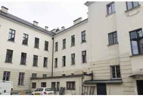 Widok wewnętrznego dziedzińca budynku szkolnego I liceum ogólnokształcącego w Cieszynie z jasną elewacją i wieloma oknami. Na dole zaparkowane pojazdy i drzwi techniczne.