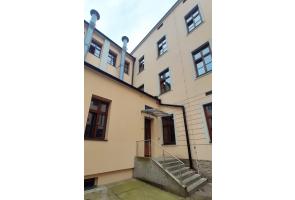 Tylna część budynku szkoły II liceum ogólnokształcącego w Cieszynie z wejściem po schodach, poręczą i zadaszeniem. Widoczne wysokie okna i jasna elewacja.