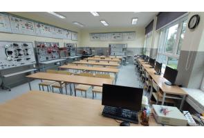 Sala dydaktyczna w Zespole Szkół Technicznych i ogólnokształcących w Skoczowie z długimi stołami i stanowiskami komputerowymi przy oknach. Na ścianach tablice edukacyjne i schematy techniczne.