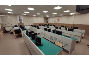 Sala komputerowa z kilkoma rzędami stanowisk wyposażonych w monitory, klawiatury i fotele. Na ścianie zegar i tablica korkowa w Zespole Szkół Technicznych w Ustroniu.