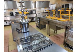Stanowisko kuchenne z płytą gazową i fontanną czekoladową. W tle zlewy, żółte krzesła na blatach i stalowe szafki kuchenne w Zespole Placówek Szkolno-Wychowawczo-Rewalidacyjnych w Cieszynie.