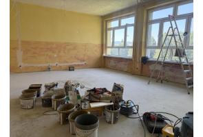 Pomieszczenie w trakcie remontu, z dużymi oknami, drabiną i odsłoniętymi przewodami pod sufitem. Na podłodze widoczne są wiadra, worki z materiałami budowlanymi, narzędzia oraz przedłużacze.