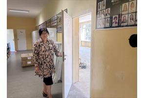 Kobieta stoi przy otwartych drzwiach prowadzących do remontowanej sali. W korytarzu widoczne są szafki, kartony i elementy wyposażenia, a w pomieszczeniu za drzwiami trwają prace przygotowawcze.