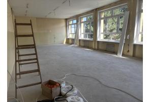 Pomieszczenie w trakcie remontu, z dużymi oknami i odsłoniętymi przewodami pod sufitem. W środku widoczna jest drewniana drabina, materiały budowlane oraz przewody leżące na podłodze.