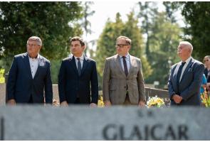 Czterech mężczyzn w garniturach stoi na cmentarzu podczas uroczystości. Na pierwszym planie rozmyta jest płyta nagrobna z napisem, a w tle widoczne są drzewa i kwiaty przy grobach.