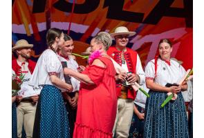 Kobieta w czerwonej sukience wręcza list gratulacyjny kobietom w strojach regionalnym. Wokół stoją pozostałe osoby w strojach regionalnych, część z kwiatami i dokumentami w dłoniach.