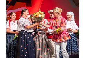 Kobieta w różowej bluzce w kwiaty przekazuje bukiet kobiecie w stroju regionalnym. Wokół stoją pozostałe osoby w strojach regionalnych, obserwujące wręczenie kwiatów.
