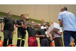 Grupa mężczyzn stoi na podwyższeniu podczas wręczania nagród. Dwóch z nich unosi złote puchary, a pozostali trzymają mniejsze trofea i nagrody rzeczowe. Przed nimi stoi mężczyzna w jasnej koszuli.