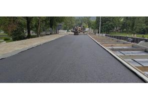 Odcinek drogi z nową nawierzchnią asfaltową podczas prac modernizacyjnych. Po bokach widoczne są nowe krawężniki i przygotowywane fragmenty chodnika, a w tle pracują maszyny drogowe.