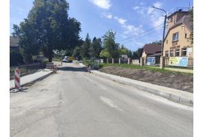 Fragment drogi podczas prac remontowych. Po obu stronach widoczne są nowe krawężniki i przygotowywane chodniki, a przy jezdni ustawiono biało-czerwone oznaczenia robót drogowych. W tle znajdują się drzewa i zabudowania.
