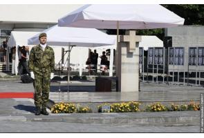 Żołnierz w mundurze stoi na placu przed pomnikiem z krzyżem. W tle widoczne są białe namioty, mikrofony oraz plansze z czarno-białymi portretami.
