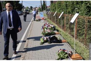 Mężczyzna w garniturze idzie wzdłuż chodnika, przy którym ustawiono tablice informacyjne. Na chodniku leżą wieńce i bukiety kwiatów, a w tle widoczne są kolejne osoby oraz pojazdy.
