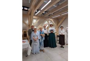 Grupa osób stoi we wnętrzu z drewnianymi belkami konstrukcyjnymi. Kilka kobiet ma stroje stylizowane na dawne, z długimi sukniami i kapeluszami, a jedna trzyma wachlarz.
