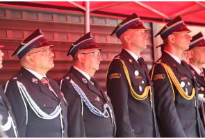 Grupa strażaków w mundurach galowych stoi w szeregu pod czerwonym zadaszeniem. Część ma ozdobne sznury i odznaczenia na mundurach, wszyscy patrzą w tym samym kierunku.