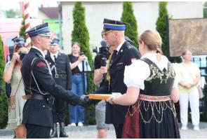Dwóch strażaków w mundurach galowych stoi naprzeciw siebie podczas wręczania dokumentu lub wyróżnienia. Obok stoi kobieta w stroju regionalnym trzymająca teczkę, a w tle widoczne są osoby obserwujące uroczystość.