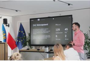 Mężczyzna w różowej koszuli stoi obok dużego ekranu i prowadzi prezentację. Na ekranie widoczny jest slajd dotyczący ZAZ Różnosprawni z danymi liczbowymi. Po lewej stronie stoją flagi a na pierwszym planie siedzą uczestnicy spotkania.