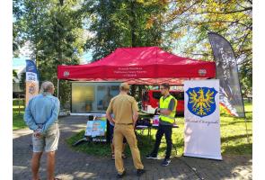 Stoisko informacyjne w parku pod czerwonym namiotem. Przed ekranem z prezentacją stoją uczestnicy oraz przedstawiciele służb, a obok znajduje się baner z herbem Powiatu Cieszyńskiego i flagi promocyjne programu OLiOC oraz Ustronia.