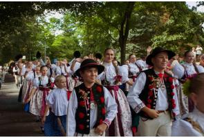 Grupa dzieci i młodzieży w strojach regionalnych uczestniczy w korowodzie, idąc alejką wśród drzew. Na pierwszym planie widoczni są chłopcy w kapeluszach i ozdobnych kamizelkach, a za nimi dziewczęta w białych bluzkach i długich spódnicach. Część uczestników macha w stronę zgromadzonych osób.