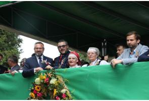 Grupa osób stoi na zadaszonej trybunie i obserwuje wydarzenie. Wśród nich są osoby w strojach regionalnych oraz eleganckich ubraniach. Z przodu trybuny widoczna jest zielona osłona ozdobiona dużym wieńcem z kwiatów, kłosów i zieleni.