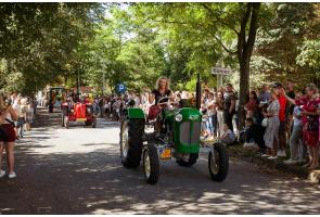 Zabytkowe ciągniki jadą w dożynkowym korowodzie alejką wśród drzew. Na pierwszym planie kobieta prowadzi zielony traktor, a za nią widoczne są kolejne pojazdy.