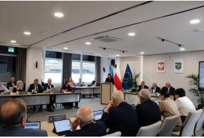Przy mównicy stoi kobieta przemawiająca do zgromadzonych, a uczestnicy siedzą przy stołach wyposażonych w mikrofony, tablety i tabliczki z nazwiskami. W tle widoczne są flagi.