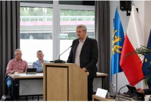 Mężczyzna przemawia przy mównicy w sali sesyjnej. Po prawej stronie widoczne są flagi Powiatu Cieszyńskiego, Polski i Unii Europejskiej, a w tle duże okna sali.