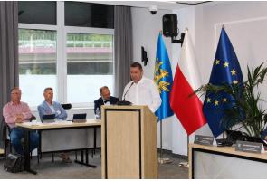 Mężczyzna przemawia przy mównicy w sali sesyjnej. Po prawej stronie widoczne są flagi Powiatu Cieszyńskiego, Polski i Unii Europejskiej, a w tle duże okna sali.