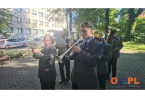 Grupa muzyków gra na instrumentach dętych podczas uroczystości plenerowej. Na pierwszym planie widoczny jest mężczyzna grający na trąbce, a za nim kolejni muzycy z instrumentami i nutami. W tle znajdują się drzewa, budynek oraz zaparkowane samochody.