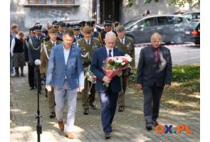 Trzech mężczyzn idzie w kierunku miejsca uroczystości, a jeden z nich niesie wiązankę biało-czerwonych kwiatów. Za nimi maszerują przedstawiciele służb mundurowych, część z nich również trzyma kwiaty. W tle widoczni są uczestnicy wydarzenia oraz zaparkowane samochody.