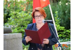 Kobieta stoi przy mikrofonie i odczytuje tekst z otwartej czerwonej teczki. W tle widoczna jest biało-czerwona flaga oraz zieleń.