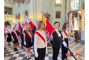 Młodzież stoi w poczcie sztandarowym we wnętrzu kościoła podczas uroczystości. Uczestnicy mają biało-czerwone szarfy i białe rękawiczki, a w tle widoczne są kolejne poczty sztandarowe oraz zgromadzone osoby.