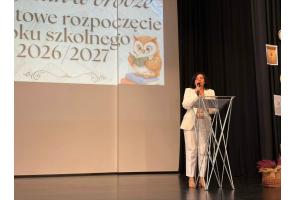 Kobieta stoi za mównicą na scenie i przemawia do mikrofonu. W tle wyświetlana jest prezentacja, a obok widoczne są czarne kotary oraz dekoracje.